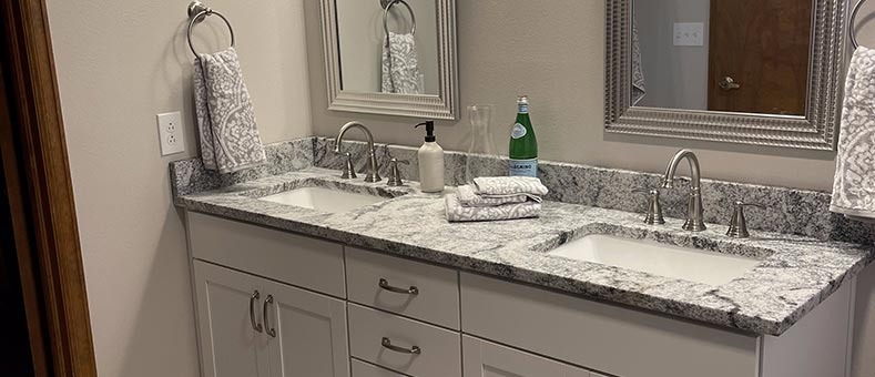 bathroom-remodeling-services A bathroom vanity with two sinks, granite countertop, silver faucets, mirrors above each sink, hand towels on rings, a soap dispenser, a bottle, and folded towels on the counter.