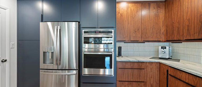 custom-carpentry-services-768×331 Modern kitchen with dark blue cabinets, stainless steel French door refrigerator, built-in oven and microwave, and wood cabinets with marble countertops and a coffee machine.