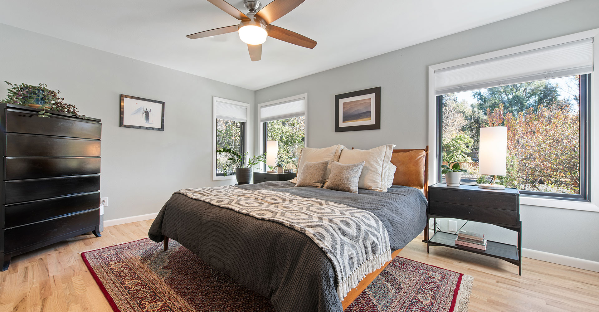 home-improvement-services Modern bedroom with a large bed, gray bedding, and patterned throw. Windows let in natural light, and there’s a dresser, nightstands with lamps, wall art, houseplants, ceiling fan, and a red area rug on hardwood floor.