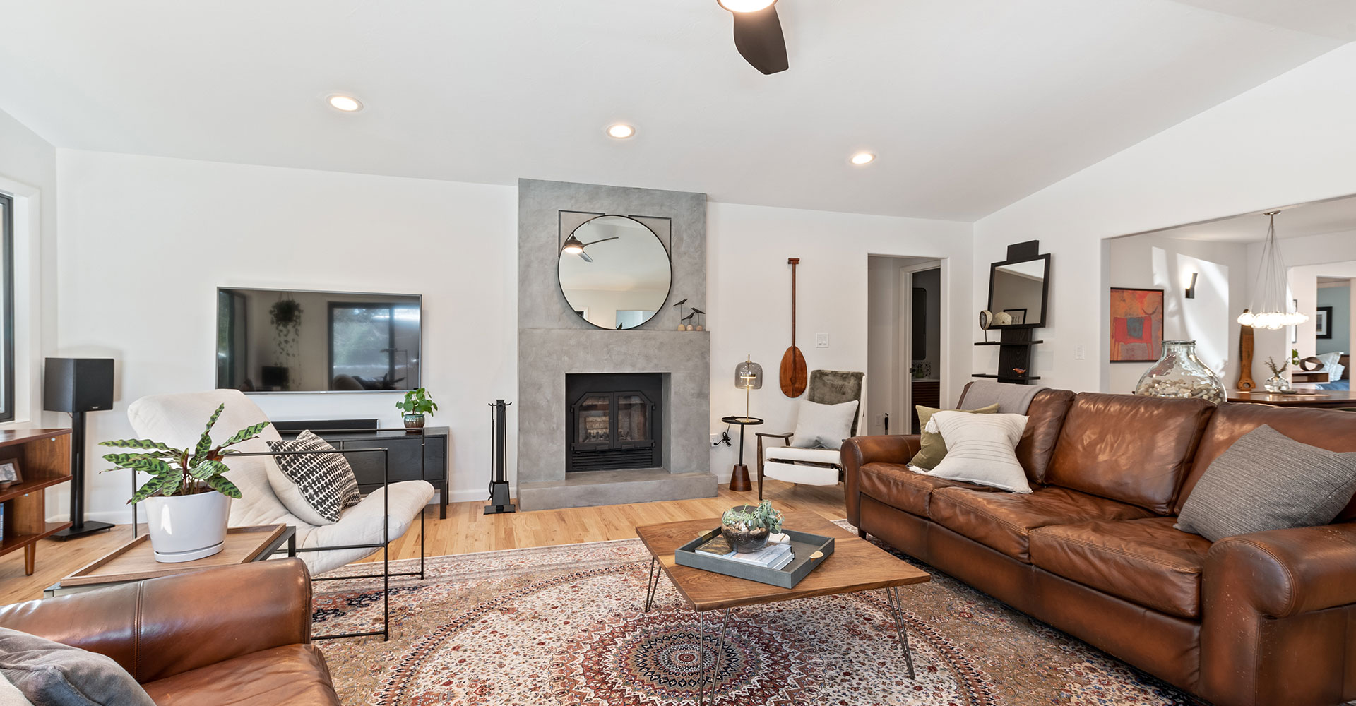 home-remodeling-services A cozy living room with a brown leather sofa, armchairs, a round rug, and a coffee table. A fireplace with a round mirror above it is centered on the back wall, with decor and plants throughout the space.