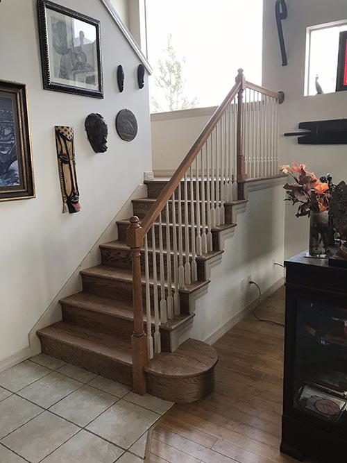 interior-carpentry-service A wooden staircase with white spindles leads up from a tiled floor to a landing. Art, masks, and framed pictures decorate the wall. A glass display cabinet with flowers sits to the right on a wooden floor.