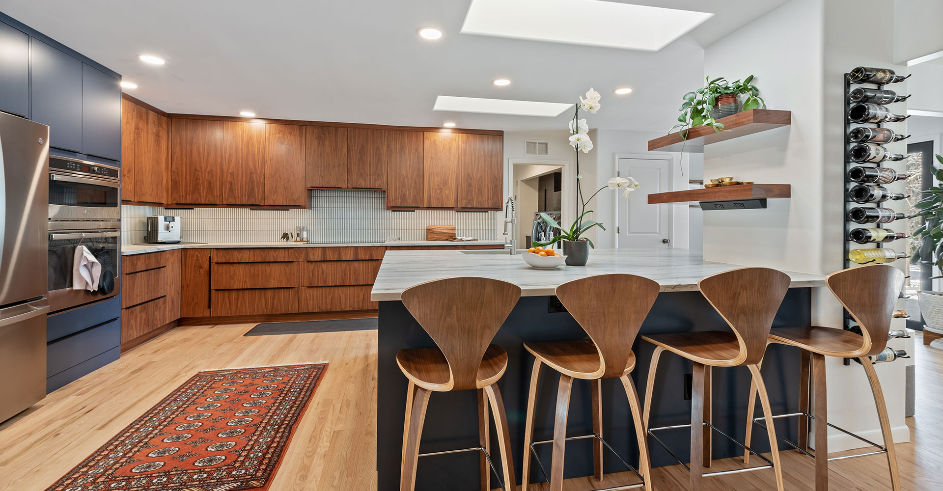 kitchen-remodeling-services Modern kitchen with wooden cabinets, stainless steel appliances, a marble island with four wooden bar stools, floating shelves with plants, a wine rack, and a patterned rug on light wood flooring.
