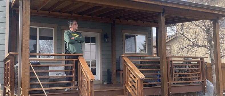 porch-building-services A man with gray hair and glasses stands with arms crossed on a wooden porch attached to a house. The porch features railings, steps, and chairs, and a leafless tree is visible in the yard.