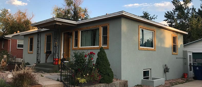 stucco-roof-installation-services A small, single-story house with light blue-gray walls and natural wood window frames, surrounded by shrubs, flowers, and a concrete walkway, under a partly cloudy sky.