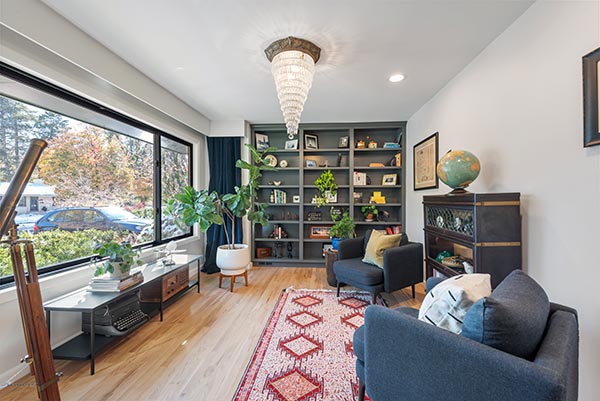 sunroom-construction-services A bright living room with large windows, a blue sofa, chairs, a red patterned rug, a chandelier, and a wall of built-in shelves filled with books, plants, and decor items. A globe sits atop a dark cabinet.