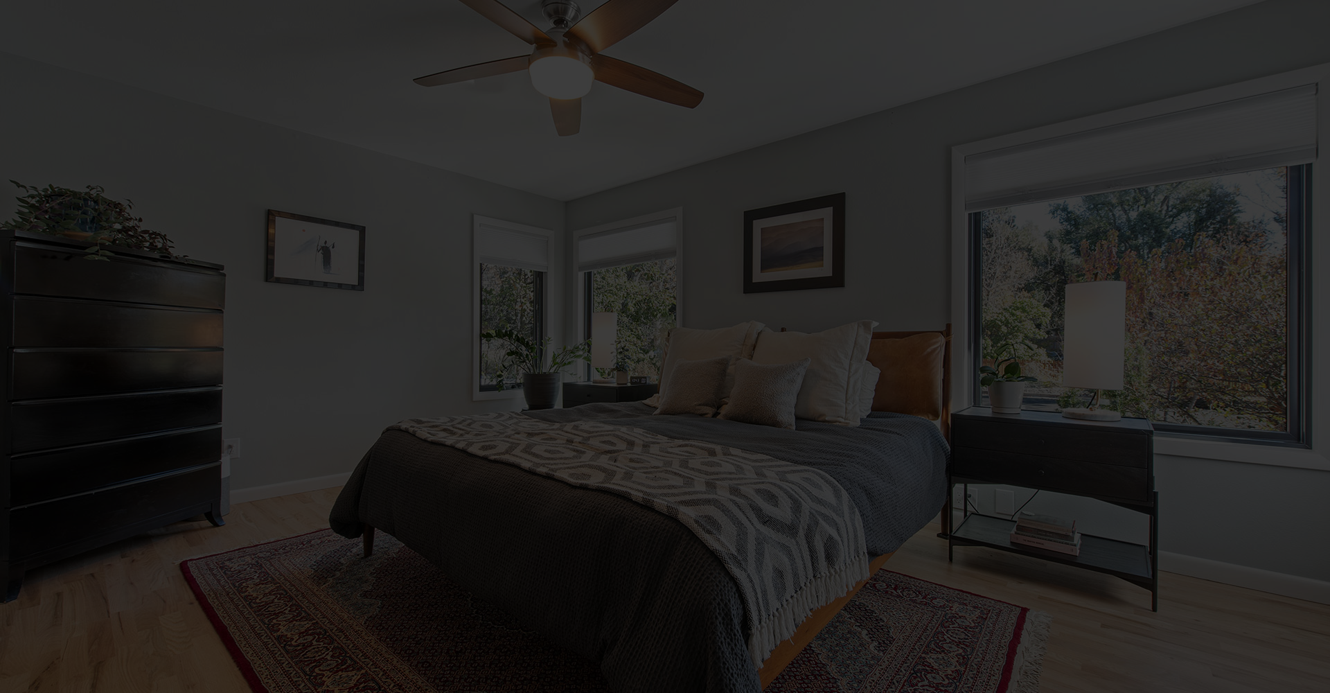 A cozy bedroom with a neatly made bed, three pillows, a dark dresser, nightstand with a lamp, framed art on light walls, ceiling fan, large windows with views of trees, and a patterned rug on the wood floor.