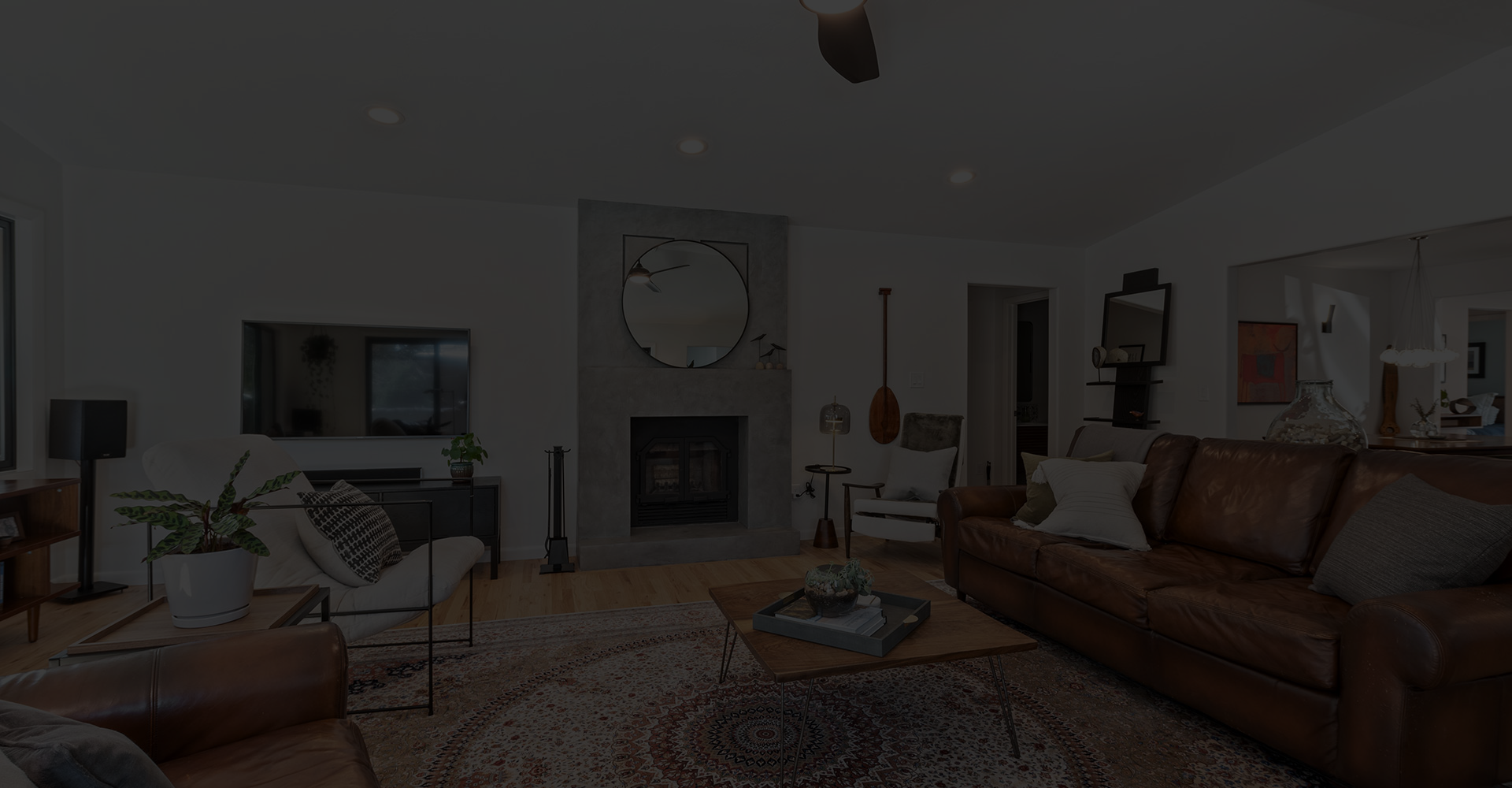 A modern living room with a brown leather sofa, patterned rug, coffee table, fireplace with a round mirror above it, wall art, plants, and neutral decor in a well-lit space.