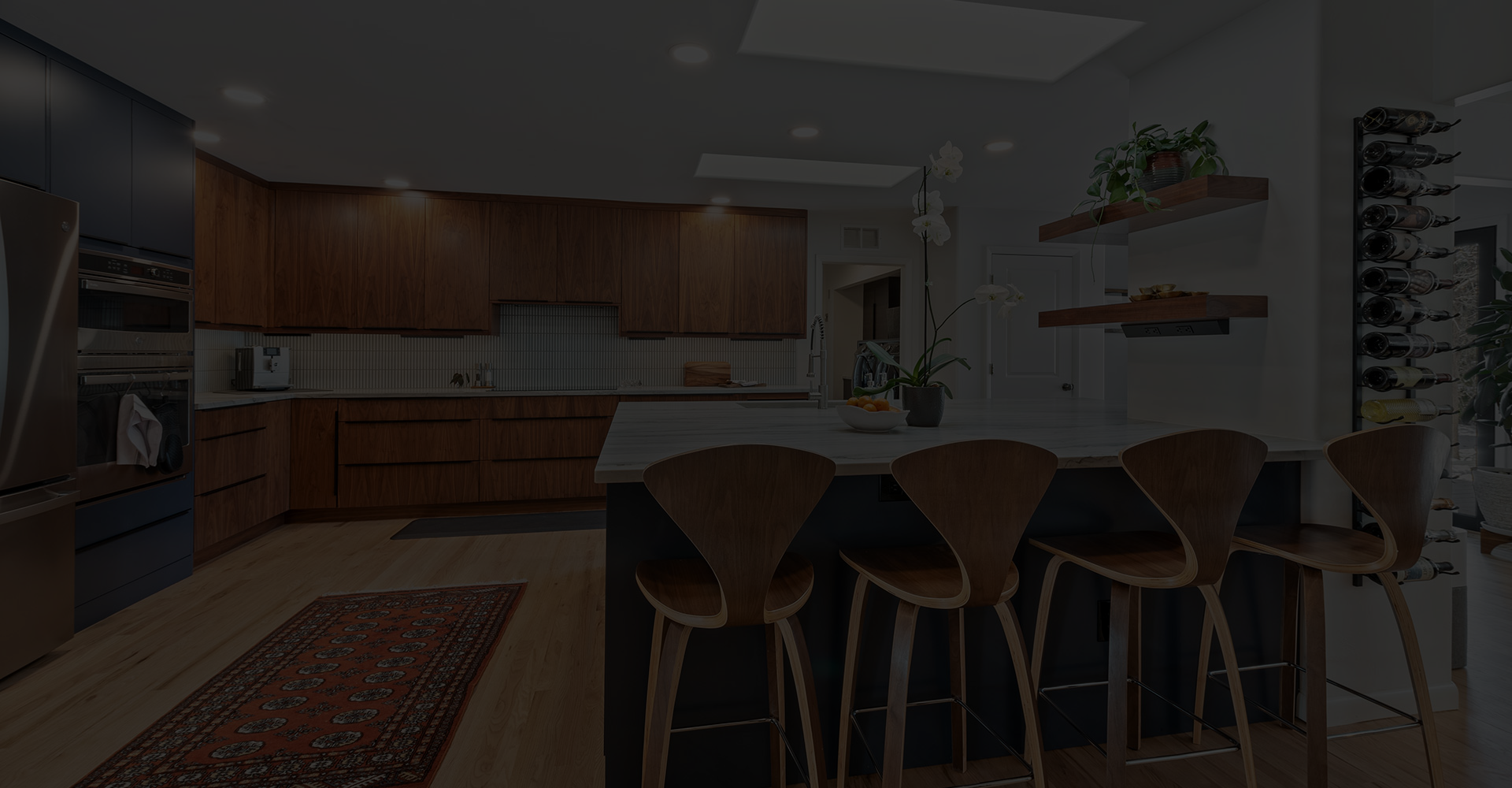 A modern kitchen with wooden cabinets, an island with four wooden bar stools, open shelves with plants, a wine rack, stainless steel appliances, and a patterned rug on a wooden floor.